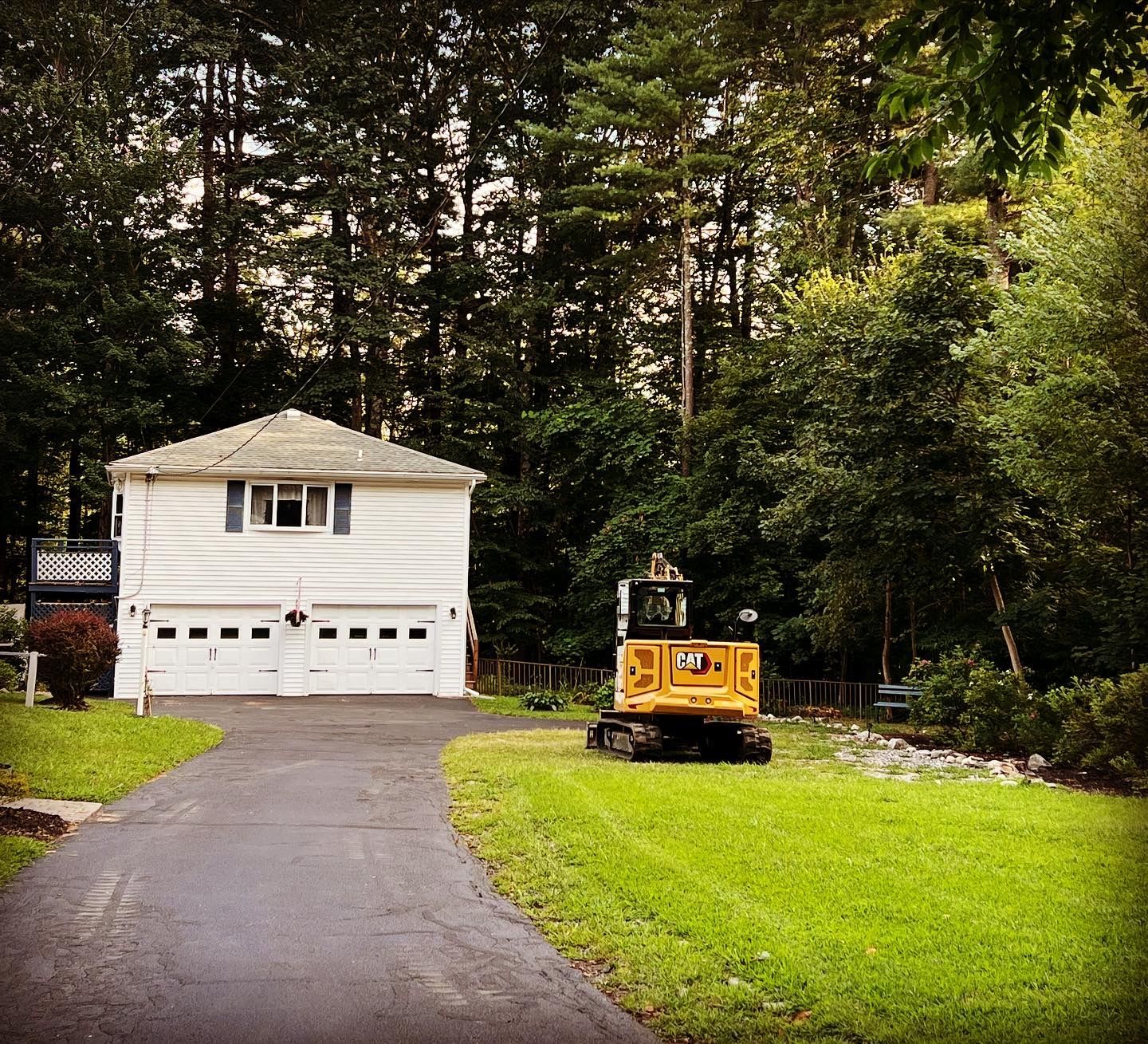 A white, two-story house with a two-car garage sits at the end of a driveway, next to a yellow construction excavator.