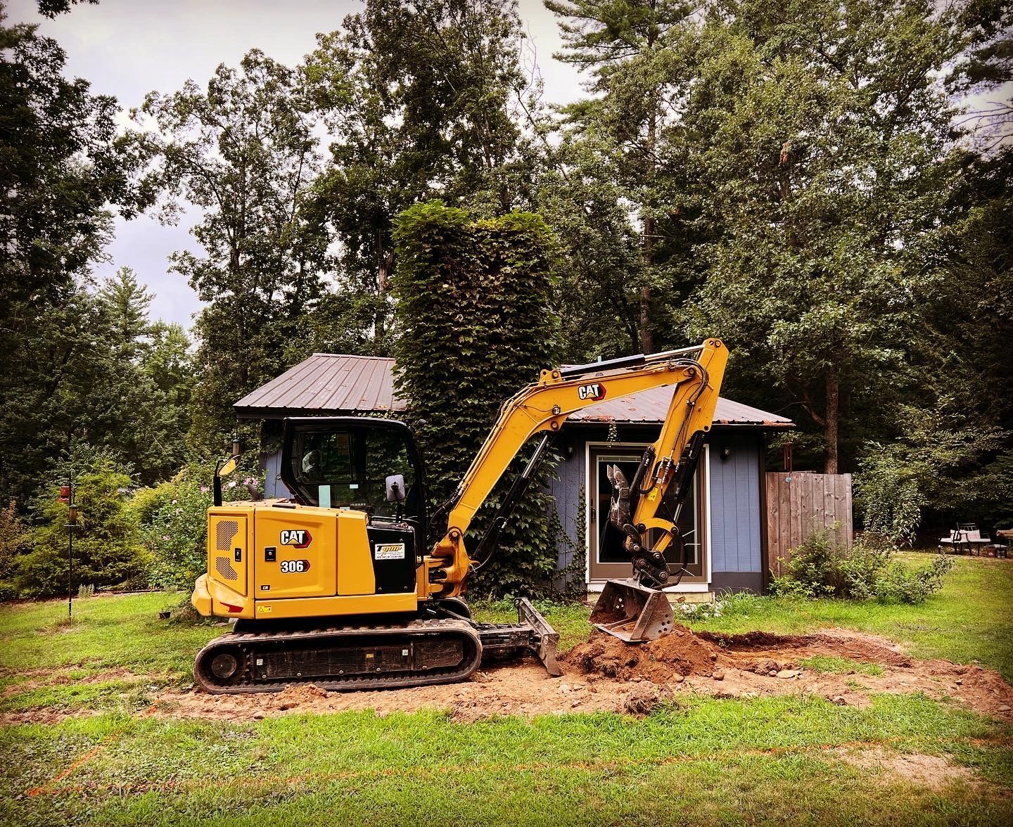 A yellow CAT mini excavator digs into the earth in front of a blue shed in a grassy, wooded area.