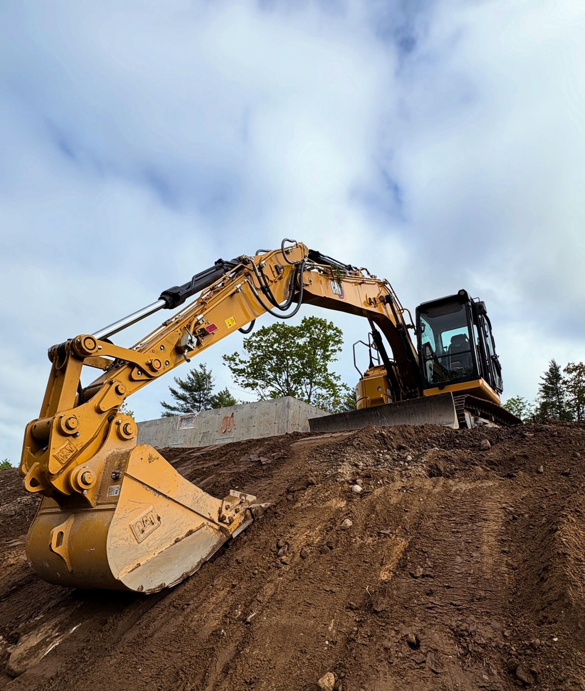 A yellow Caterpillar excavator sits on a dirt mound against a cloudy sky.