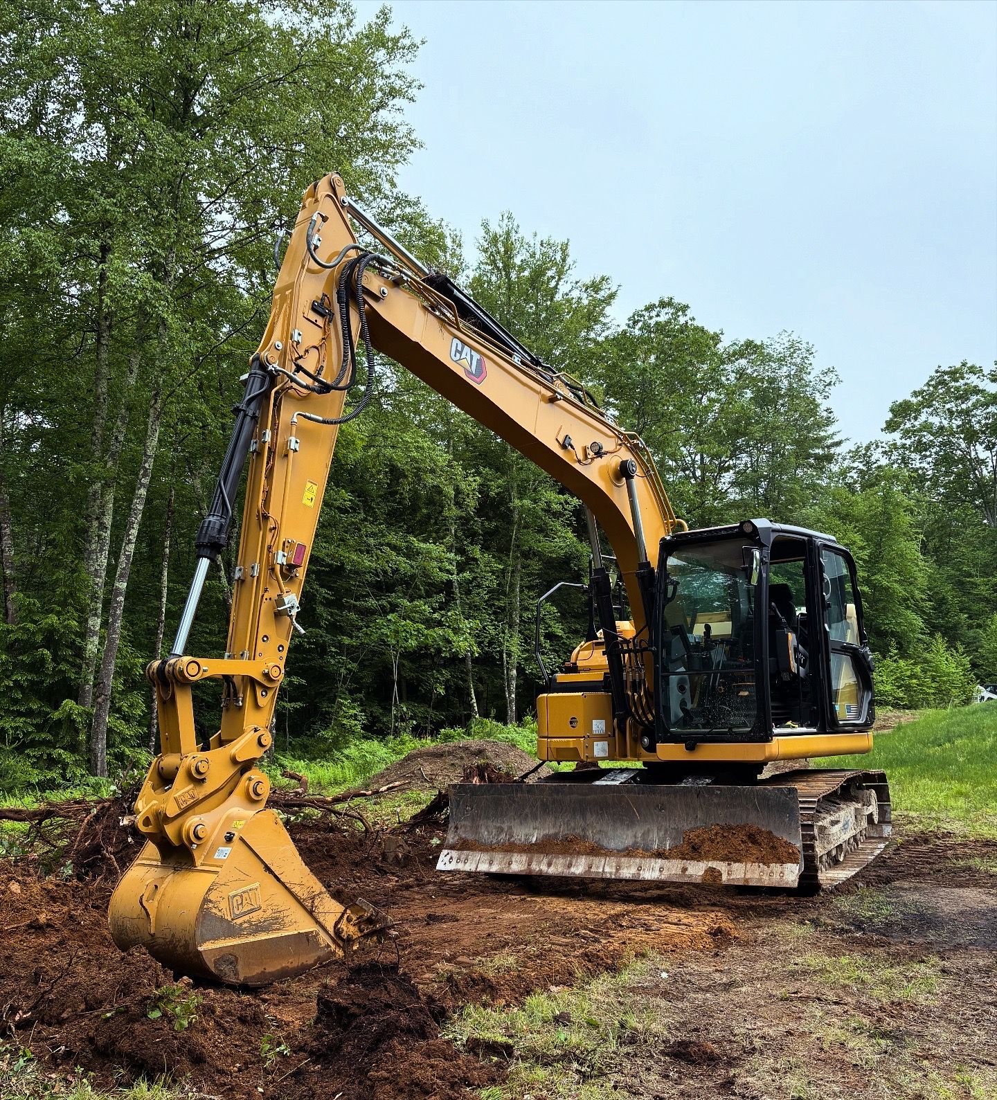A yellow Caterpillar excavator sits on a patch of loose dirt in a wooded area with its bucket lowered to the ground.