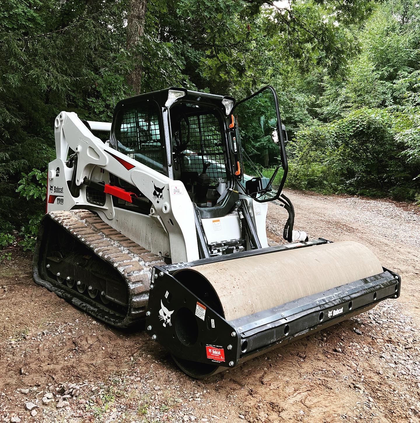 A white and black Bobcat compact track loader equipped with a large soil roller attachment parked on a gravel path.