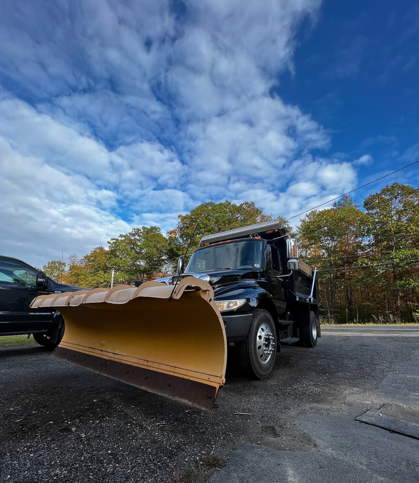 A black dump truck with a large yellow snow plow attached to the front, parked on an asphalt lot under a cloudy sky.