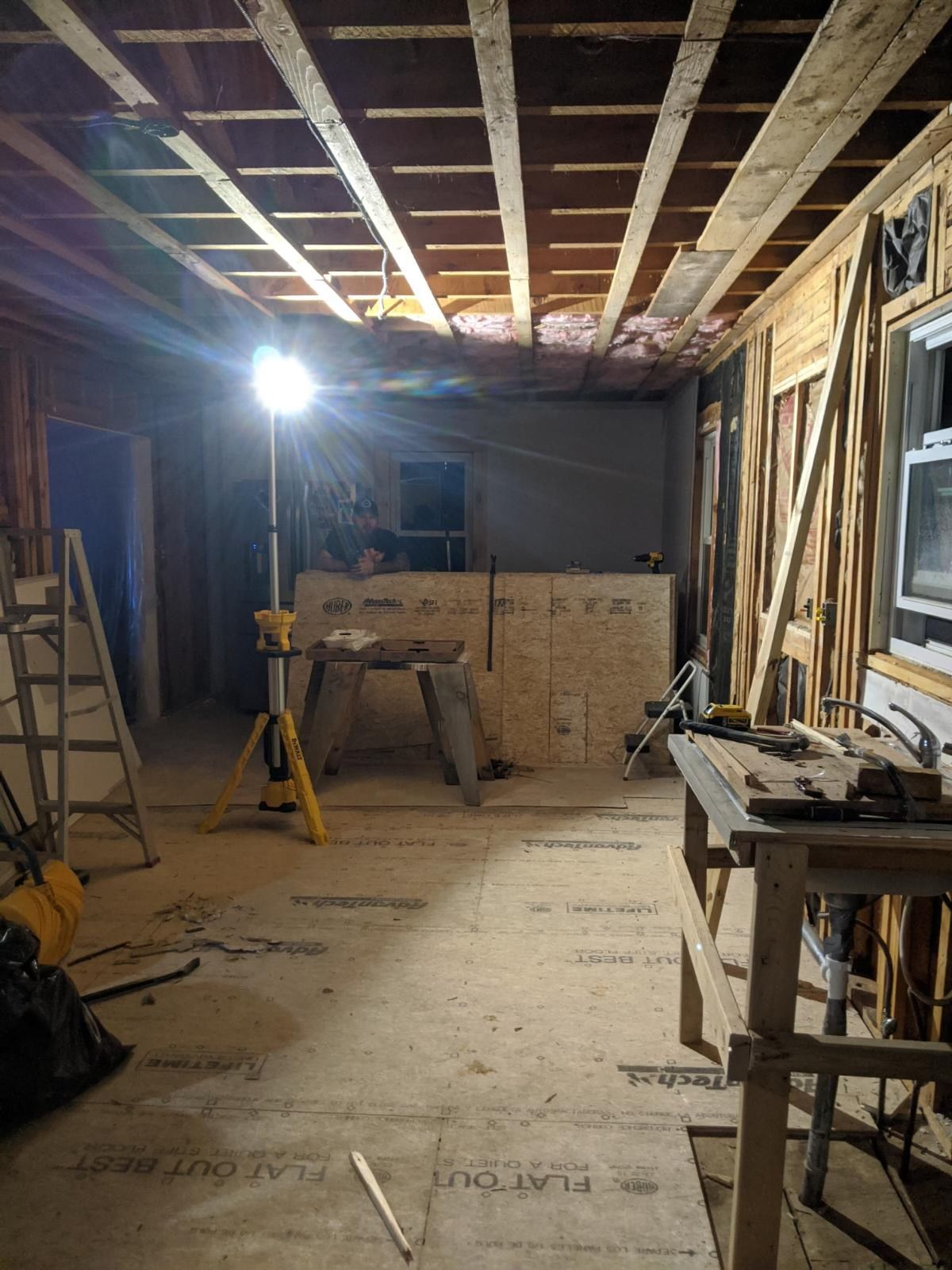 An indoor construction site with a tripod work light, exposed ceiling beams, and plywood floor underlayment.