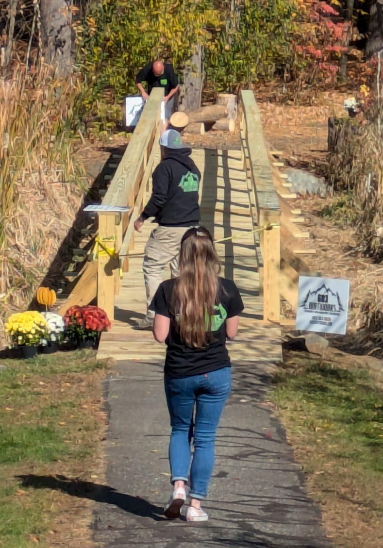 Three people work on constructing a wooden footbridge outdoors, surrounded by autumn foliage and potted chrysanthemums.