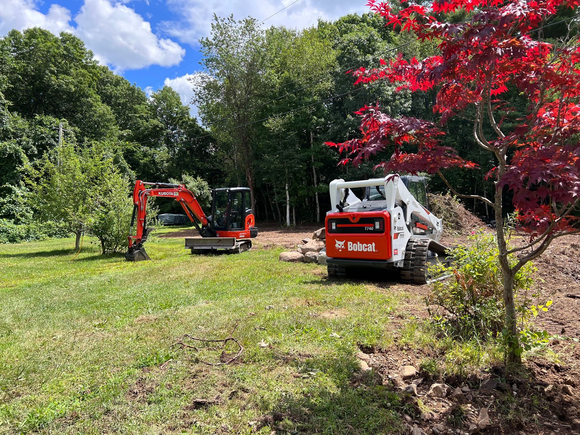 An orange excavator and a white Bobcat skid-steer sit in a grassy, partially cleared field near a forest and a red tree.