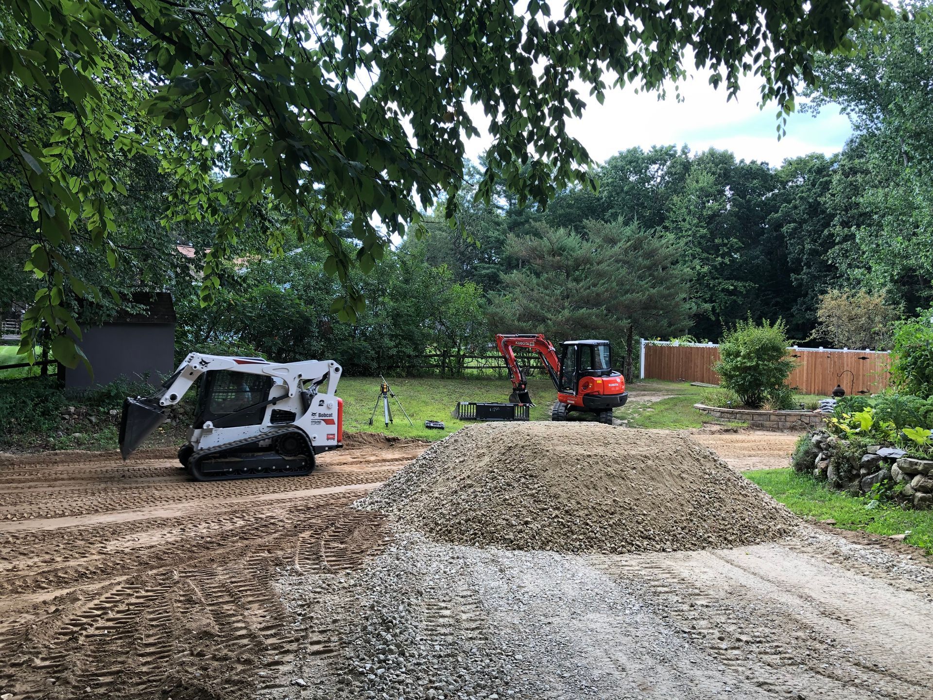A skid steer and an orange excavator sit on a dirt lot with a large pile of gravel in the foreground.
