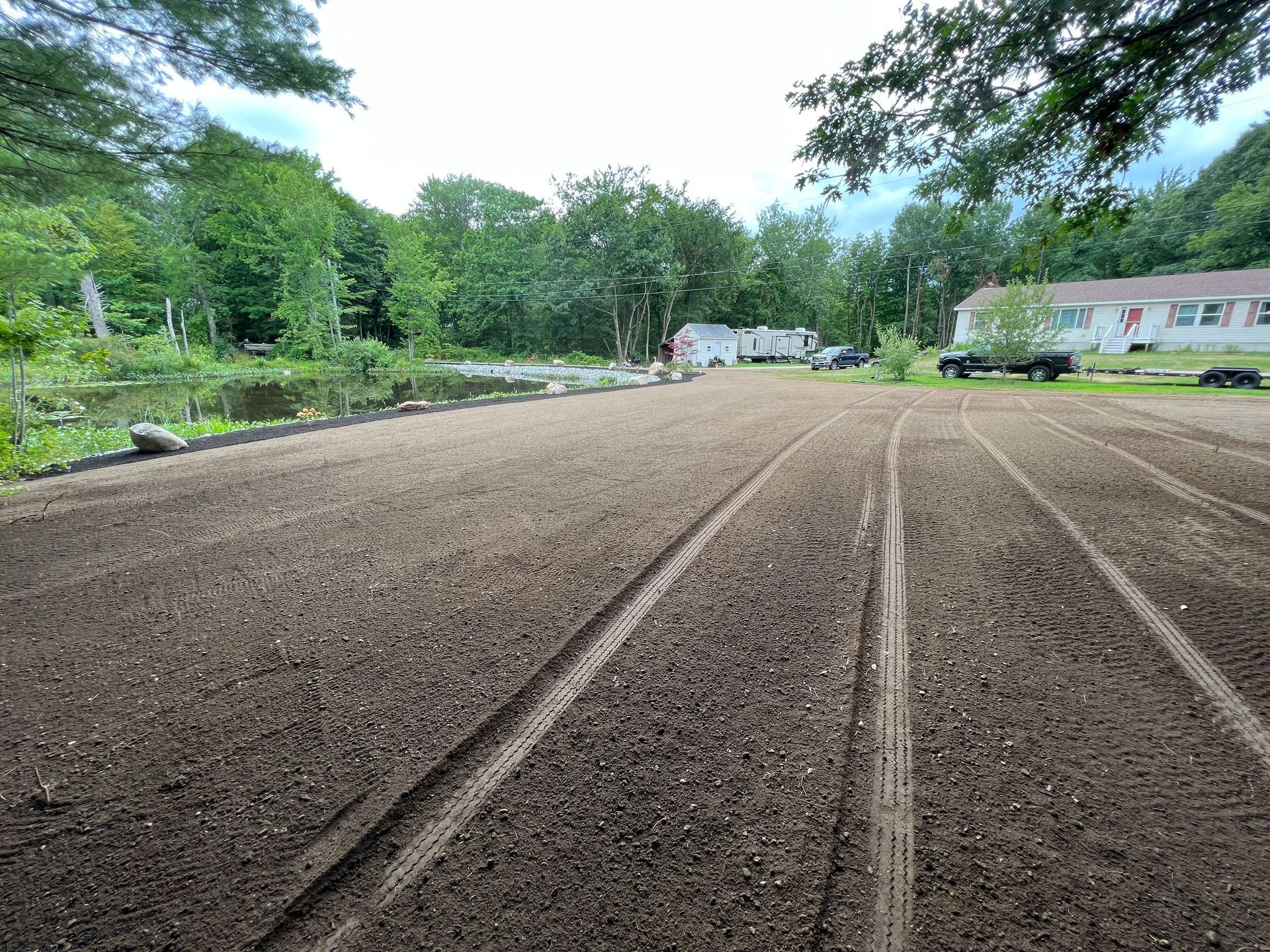 Freshly tilled dirt yard with tire tracks leading toward a pond, a house, and a camper in a rural setting.