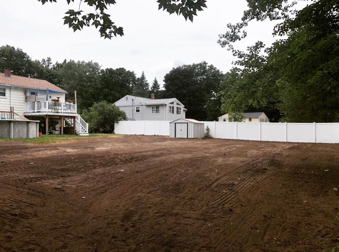 A large, cleared dirt backyard with a white vinyl fence, a shed, and neighboring houses in the background.