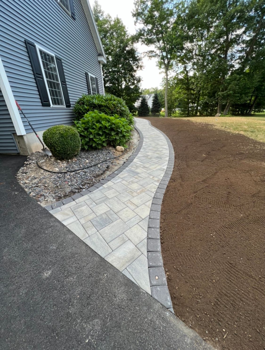 A curved stone walkway leads from an asphalt driveway toward a blue house, flanked by manicured shrubs and fresh topsoil.