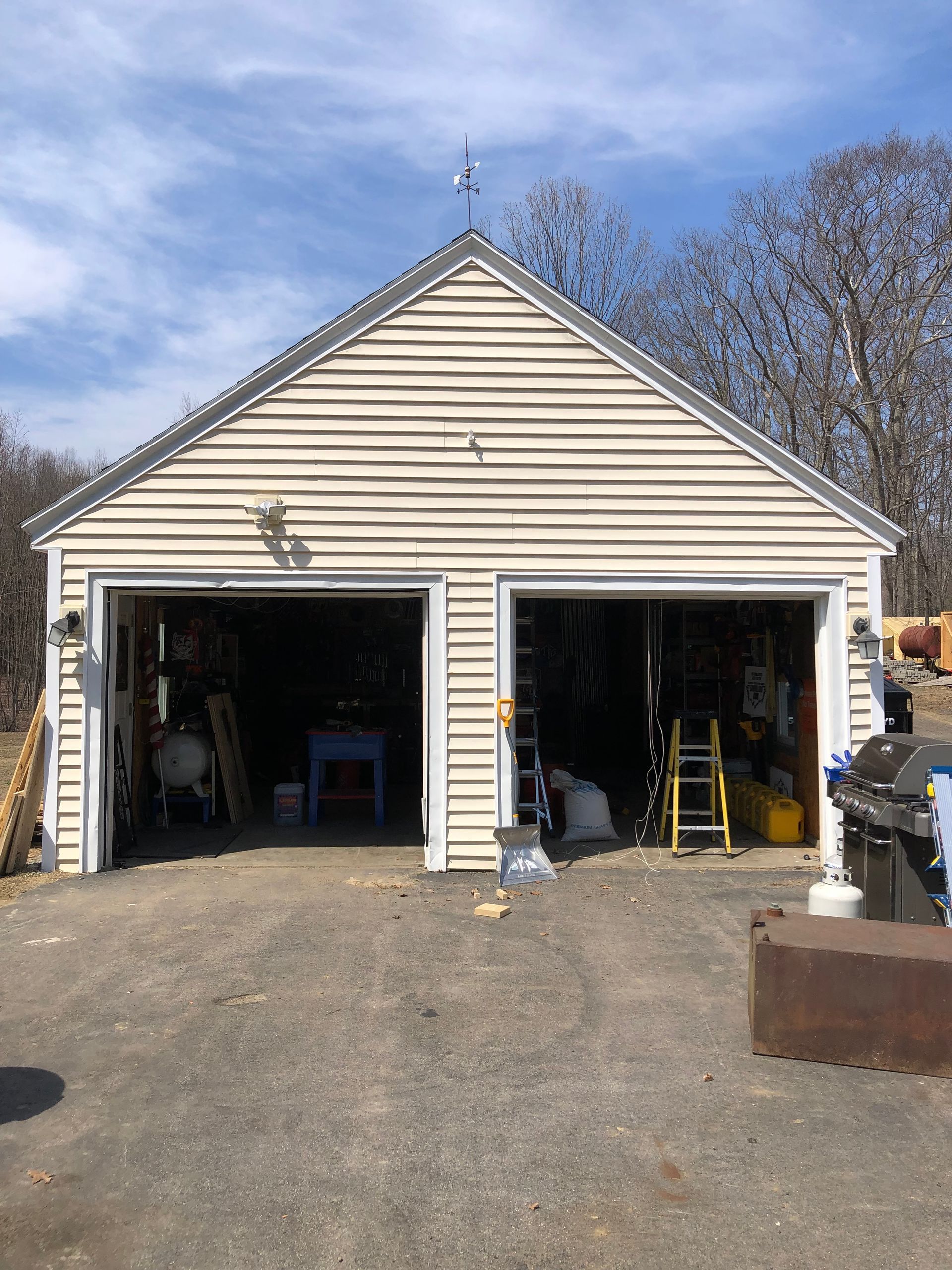 A detached, light-colored vinyl-sided garage with two open bays, standing on a gravel driveway under a blue sky.