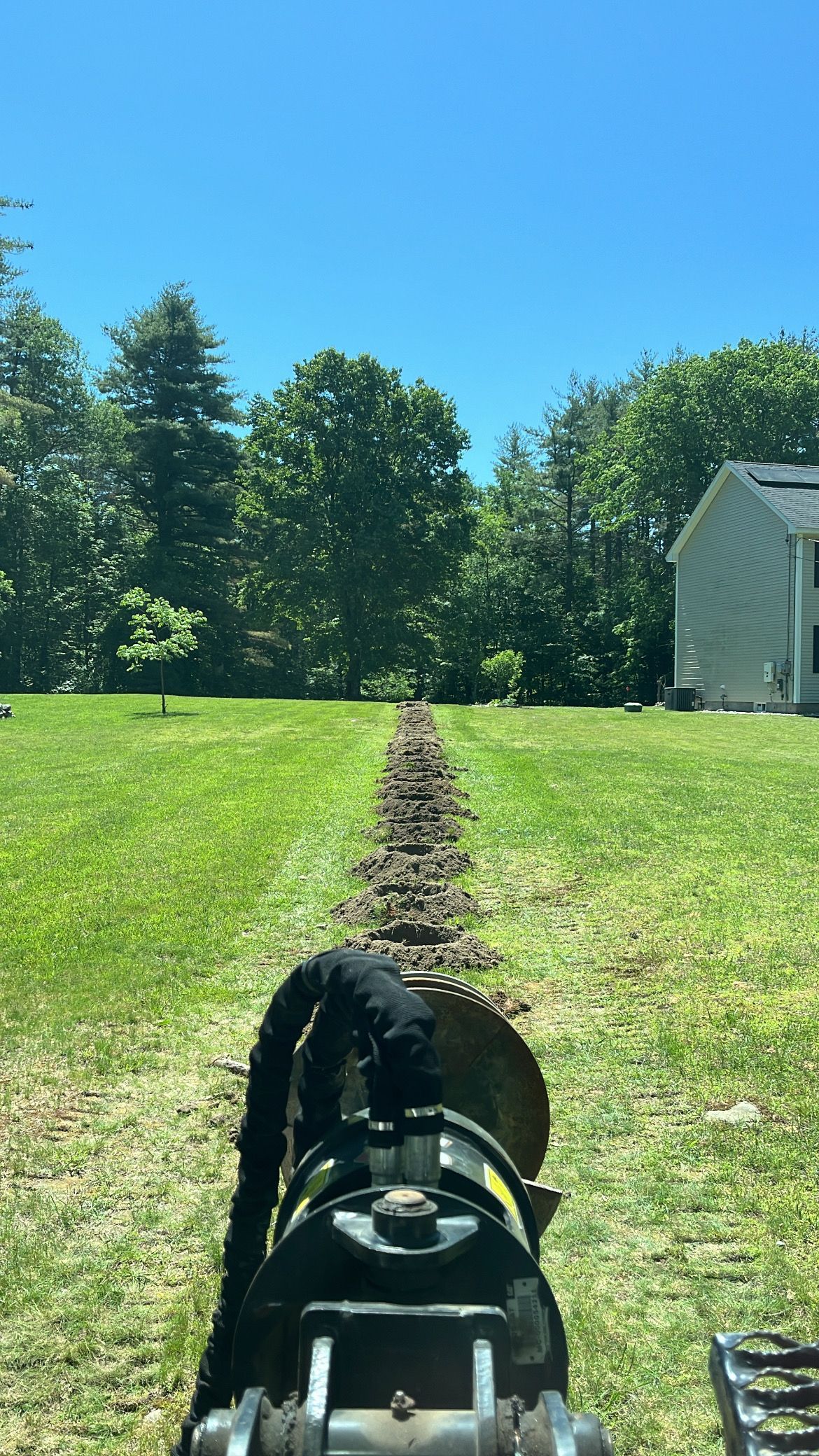 A view from a tractor looking down a freshly dug trench through a grassy yard toward a house in the distance.