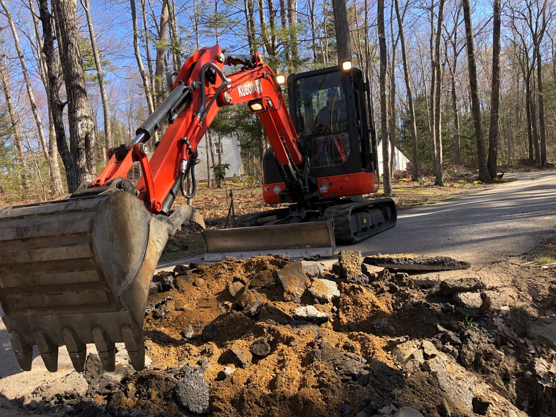 A bright orange excavator sits on a dirt path, digging into a mound of earth and rocks in a wooded area.