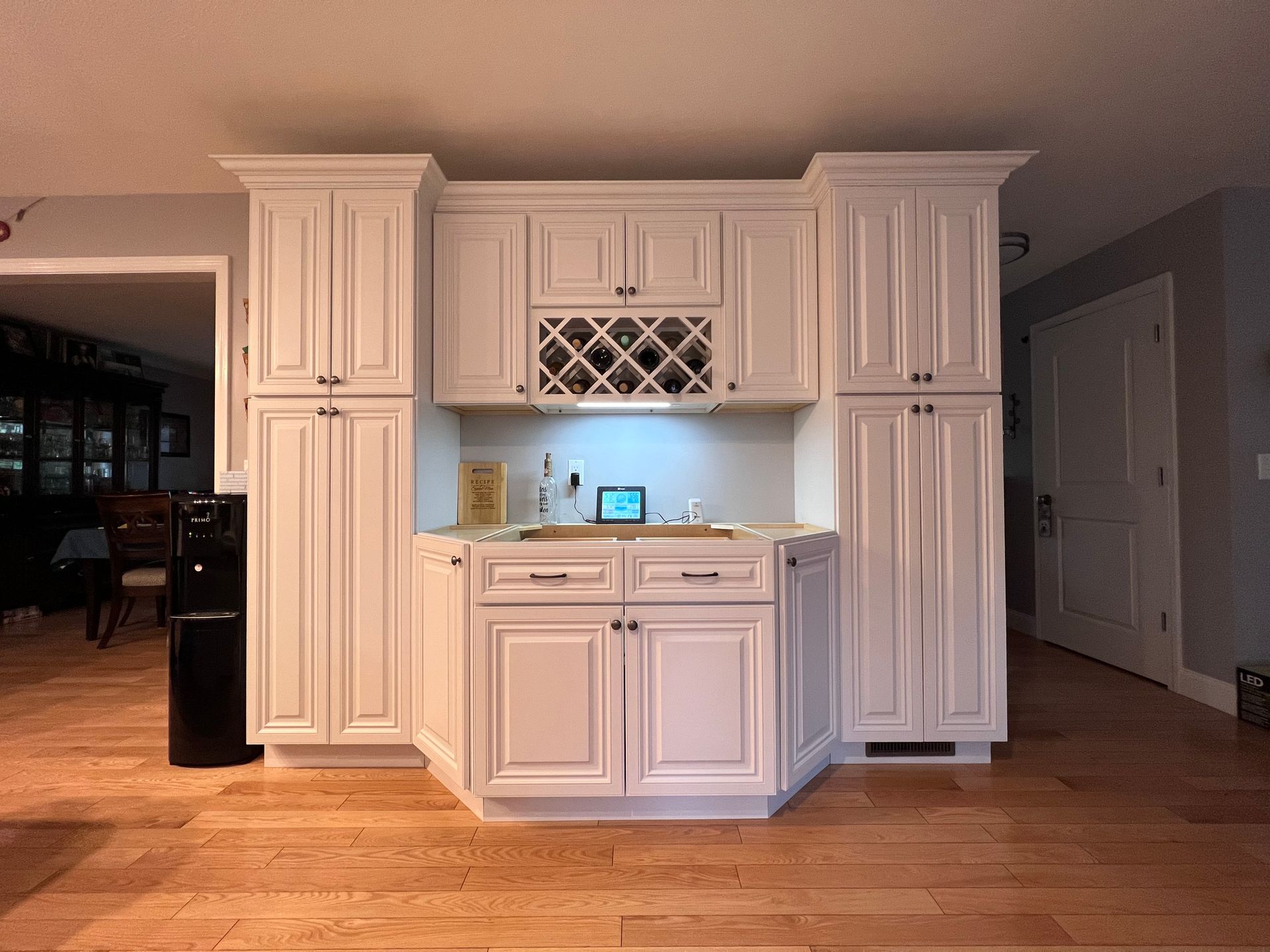 White floor-to-ceiling cabinets framing a central wet bar with a wine rack and a counter on light hardwood flooring.