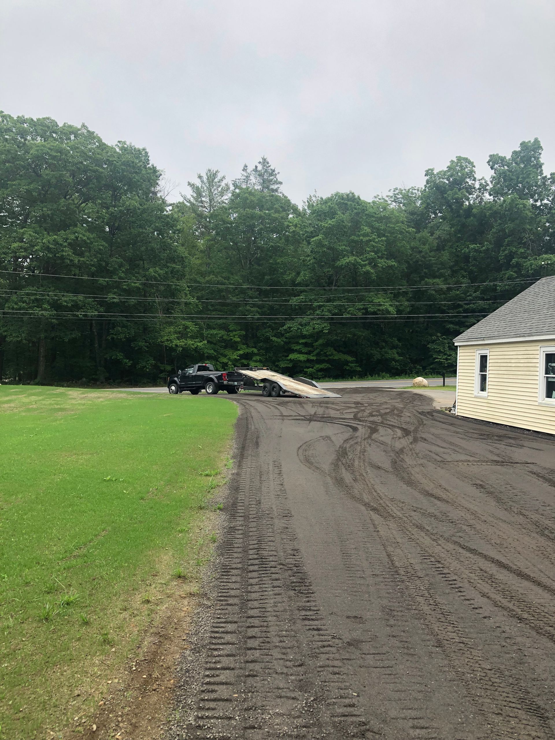 A dark pickup truck towing an empty flatbed trailer on a muddy driveway next to a grassy field and a light-colored building.