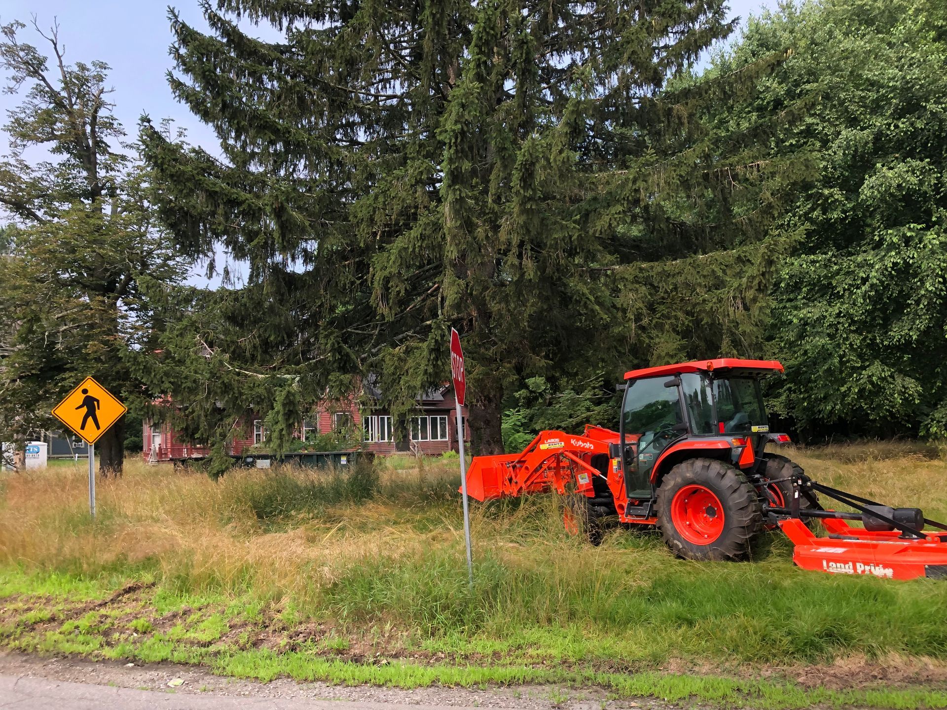 A bright orange tractor with a mower attachment is parked in a grassy field next to a yellow pedestrian crossing sign.