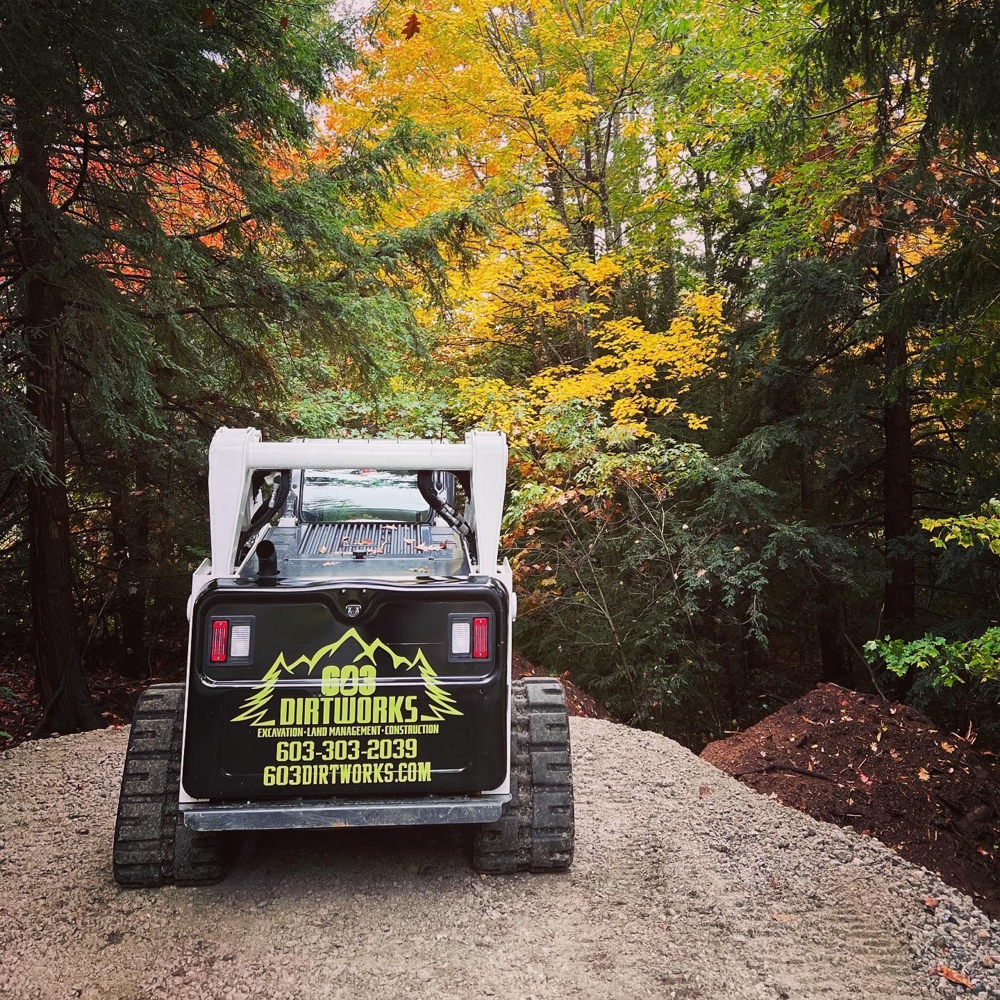 A white tracked skid steer parked on a gravel path in a forest with autumn-colored trees, displaying company branding.