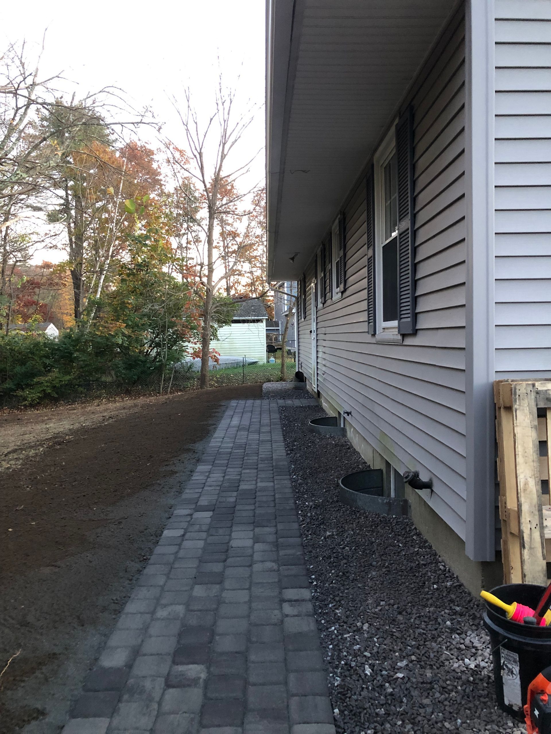 A paved stone walkway runs along the side of a house with grey siding and a gravel border, with trees in the background.