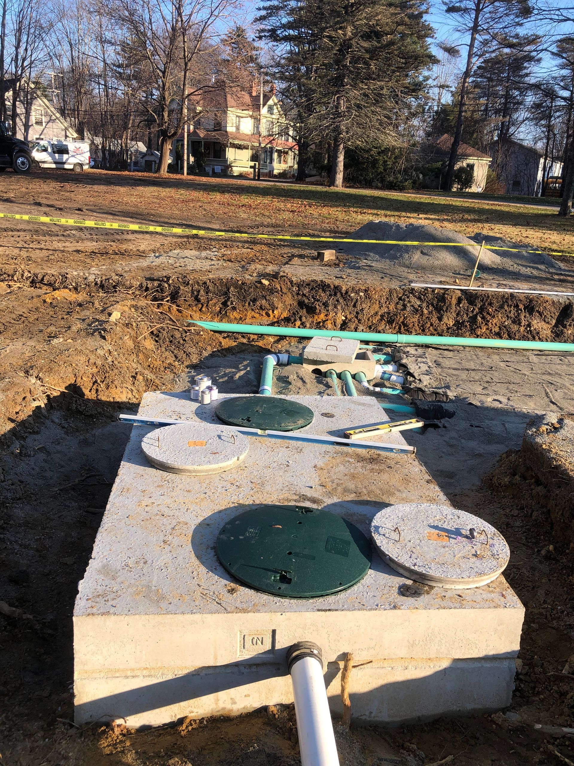 A concrete septic tank with green covers sits in an excavated trench outdoors, connected to white and green pipes.