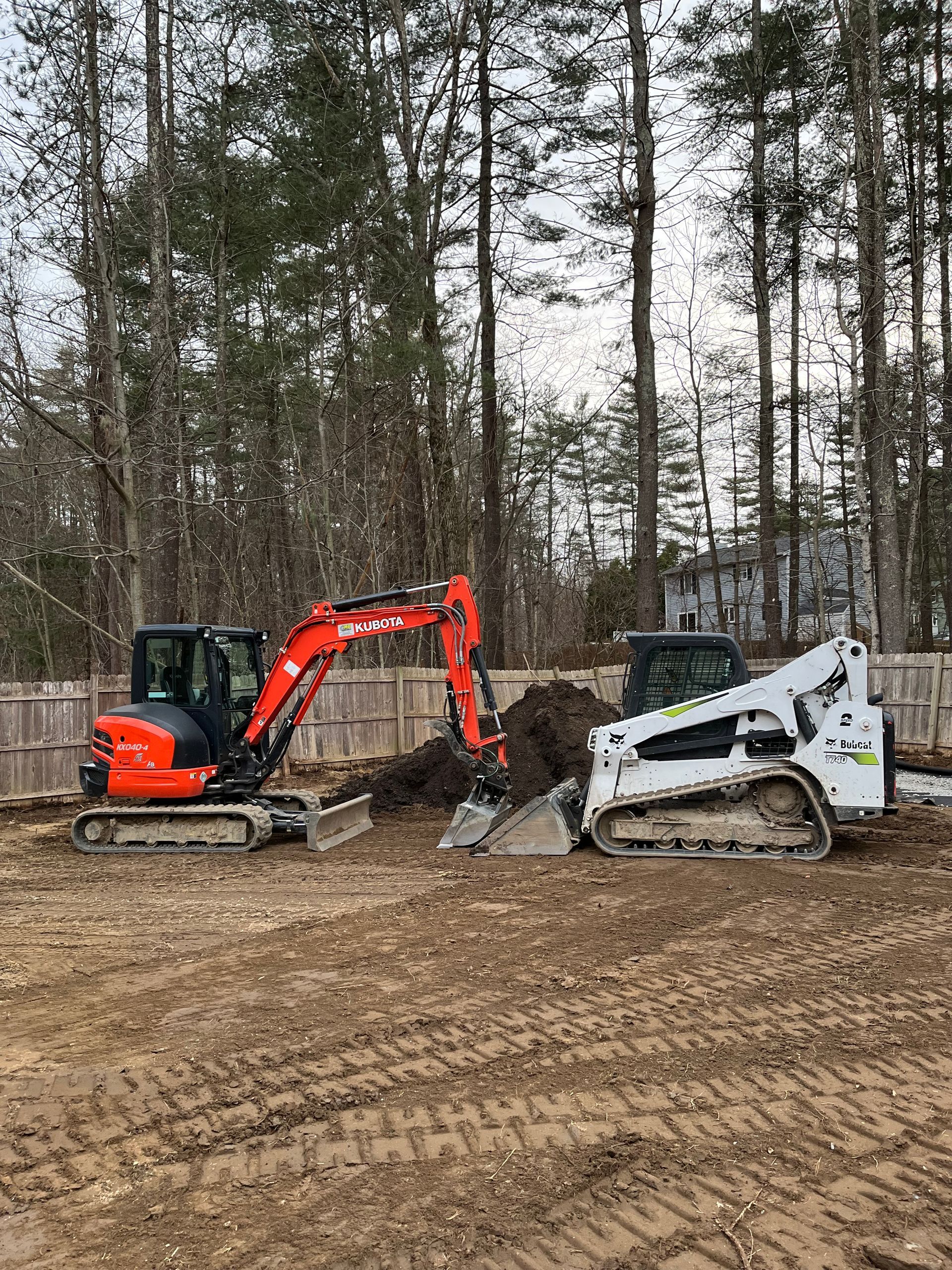 An orange Kubota excavator and a white Bobcat skid steer working together on a dirt lot in front of a tree line.