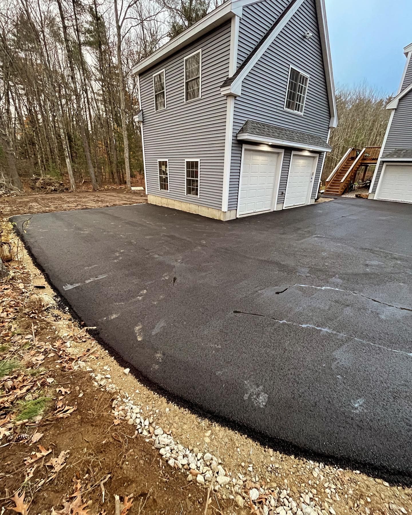 A newly paved asphalt driveway leads to a two-story gray house with two garage doors and a gravel border.