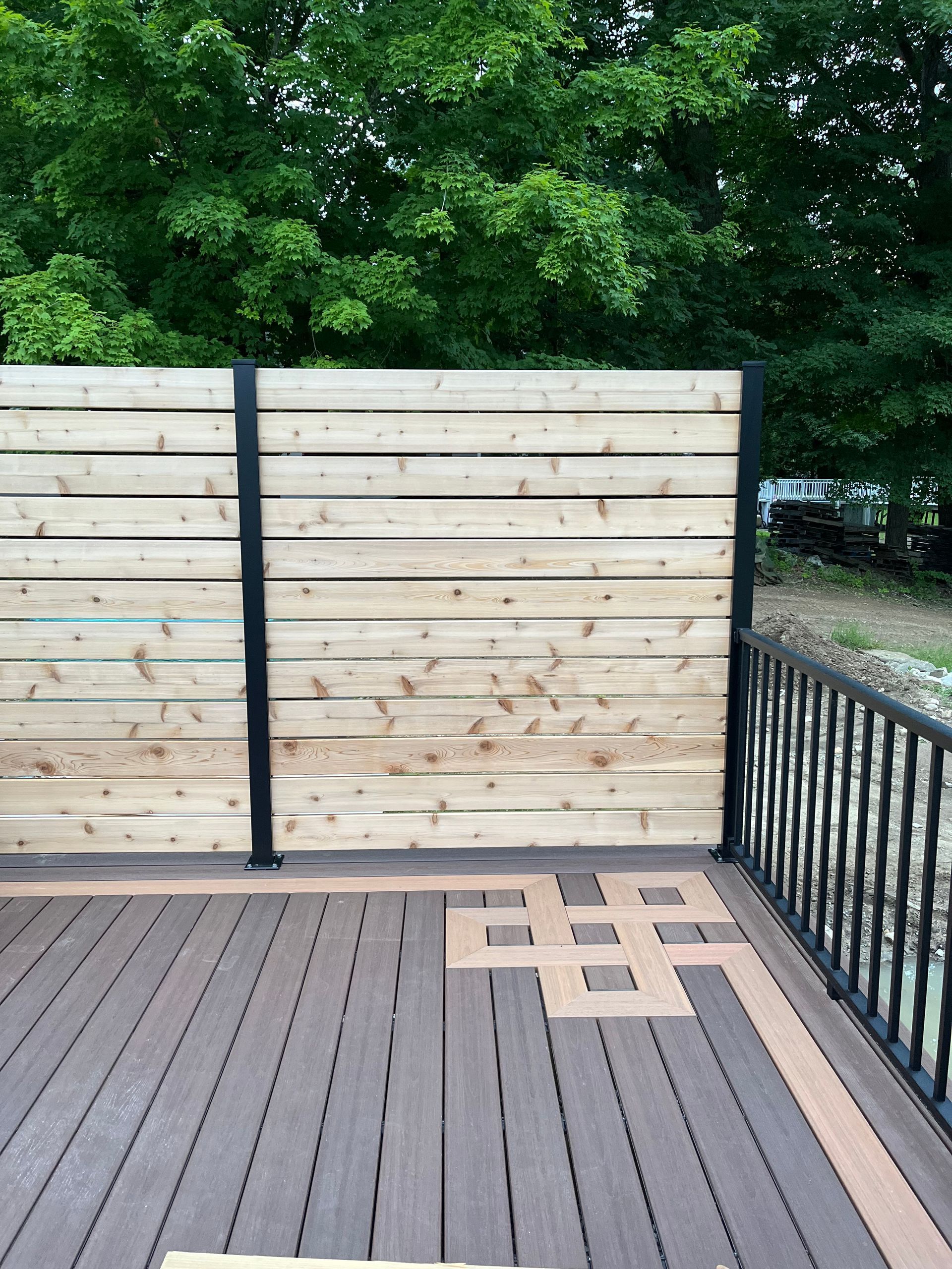 A wooden horizontal slat privacy screen stands on a composite deck with a decorative inlay pattern and black railings.