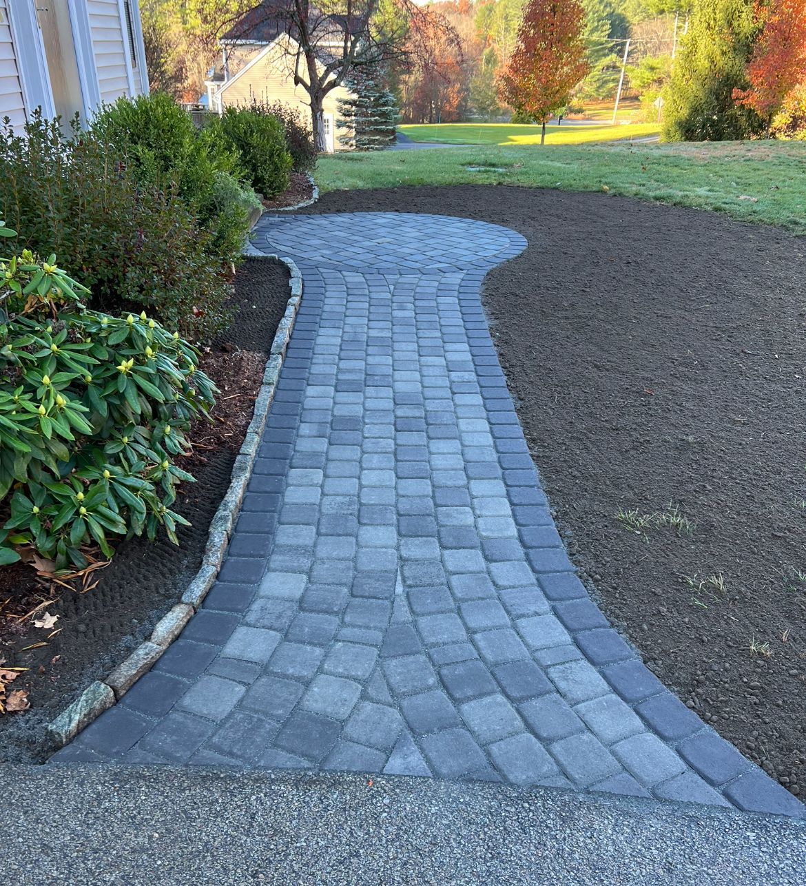 A grey stone paver walkway leads from a patio through a yard toward a house surrounded by trees in the background.