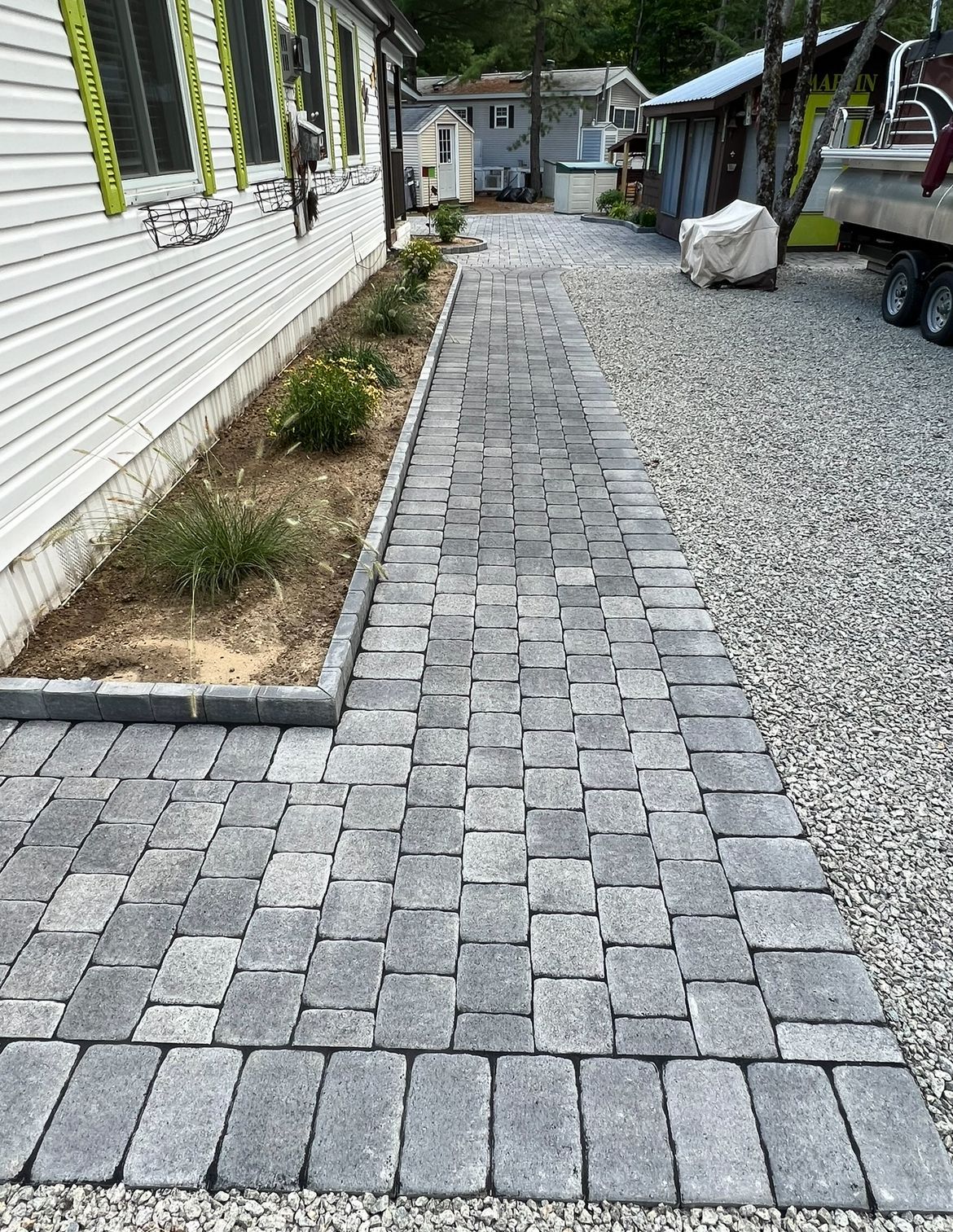 A new gray paver walkway runs alongside a white building with a landscaped border and a gravel driveway on the right.
