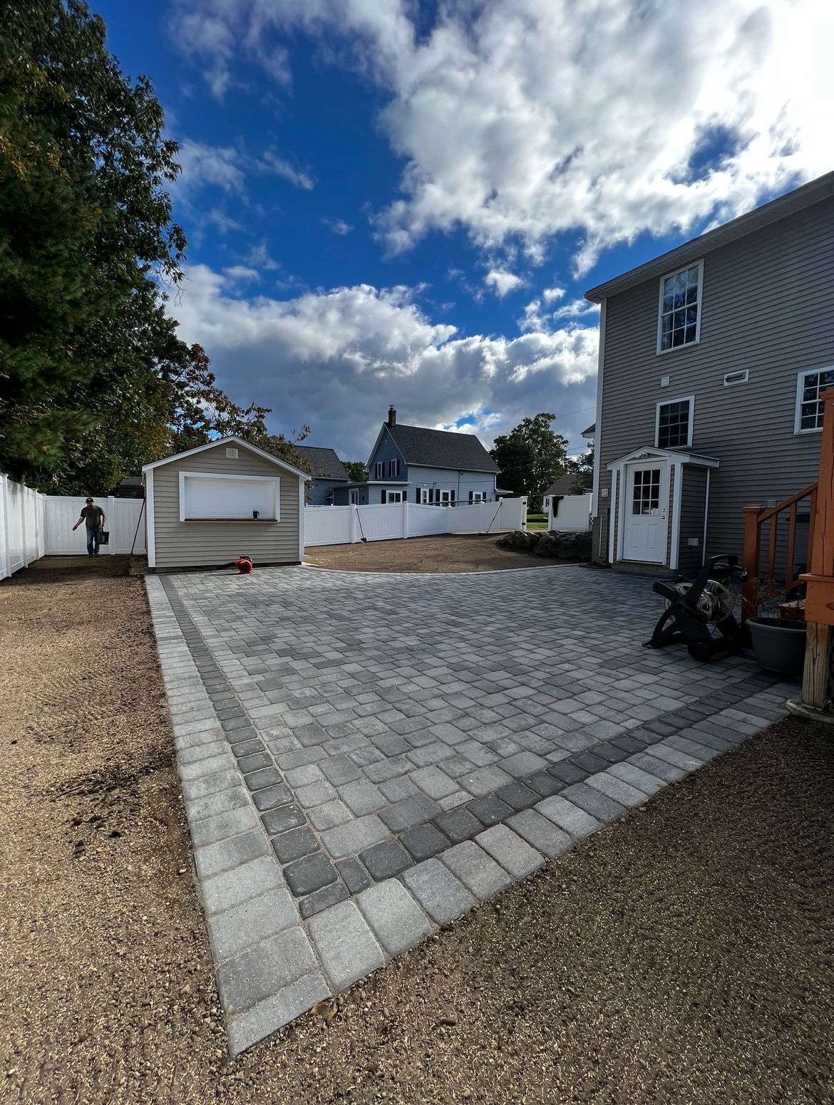 A new gray paver patio with a dark border sits in a backyard next to a house and shed under a partly cloudy blue sky.