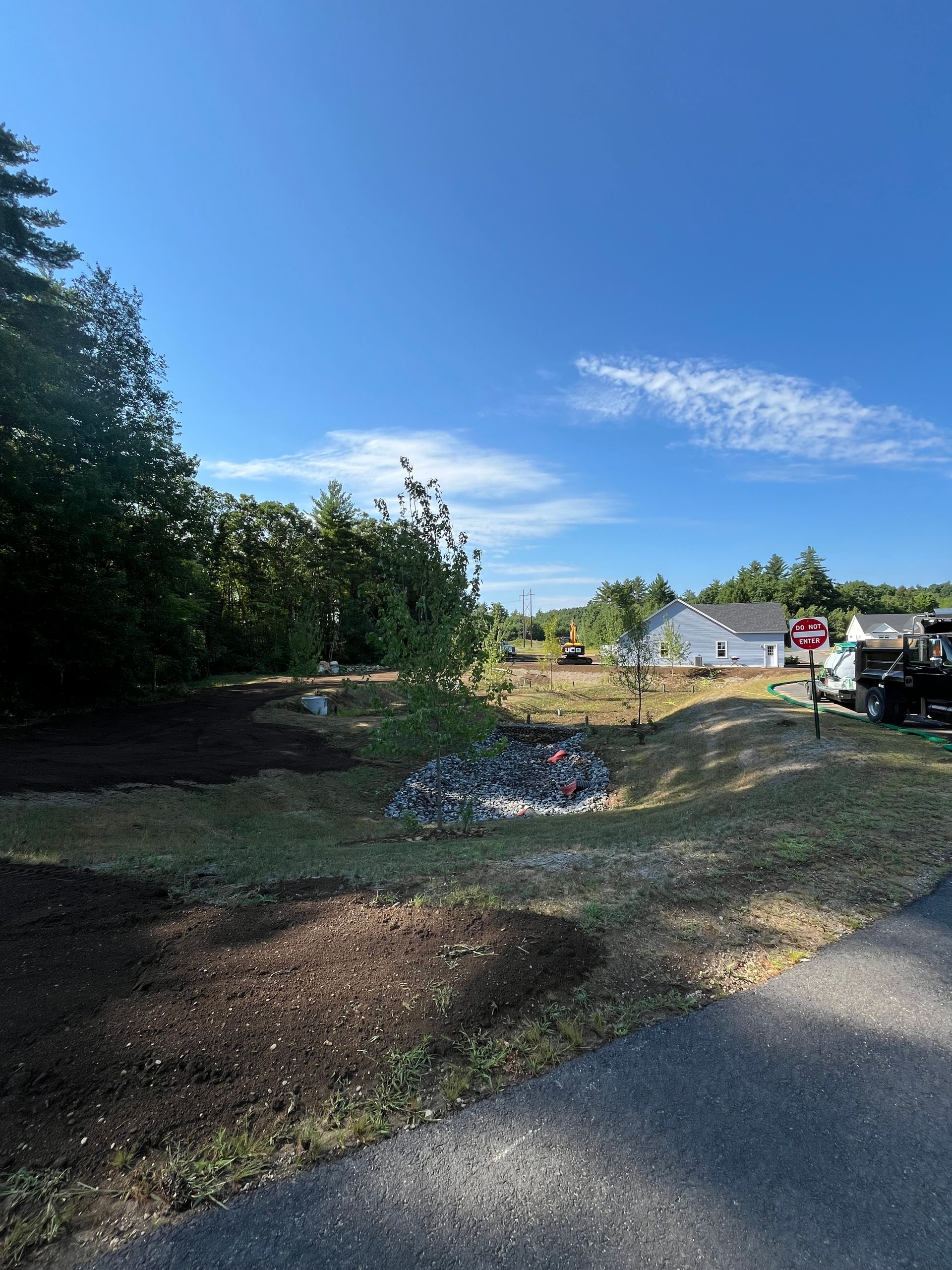 A landscaped drainage area with mulch, rocks, and grass under a clear blue sky, next to a paved path and distant houses.