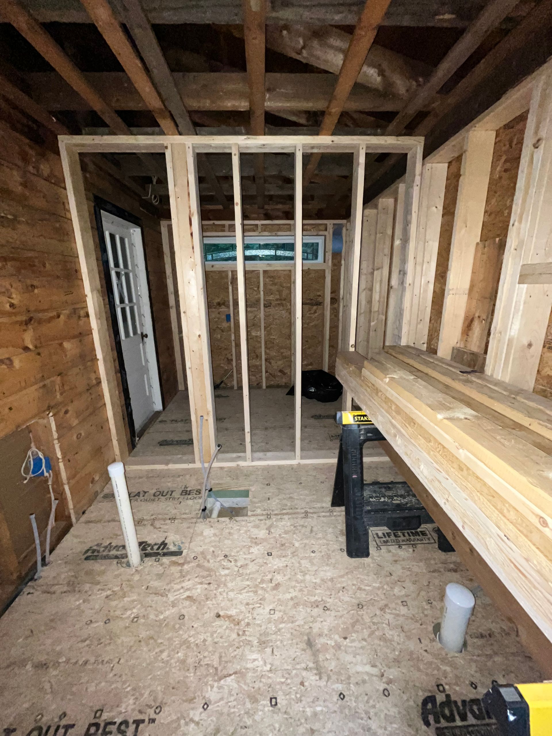 Framed interior wall in a room under construction, with exposed wooden studs, a white door, and subflooring visible.