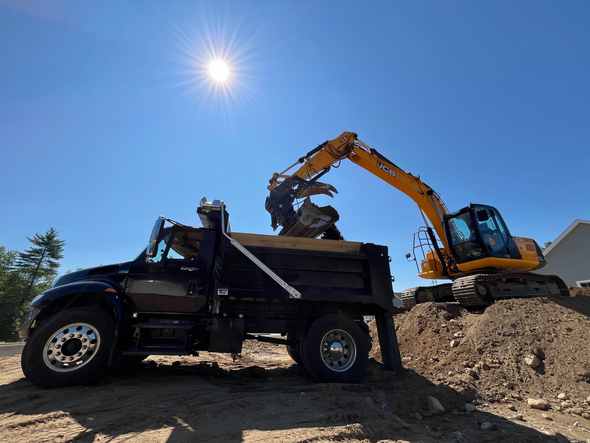 A yellow excavator loads dirt into the bed of a black dump truck against a clear, sunny sky.