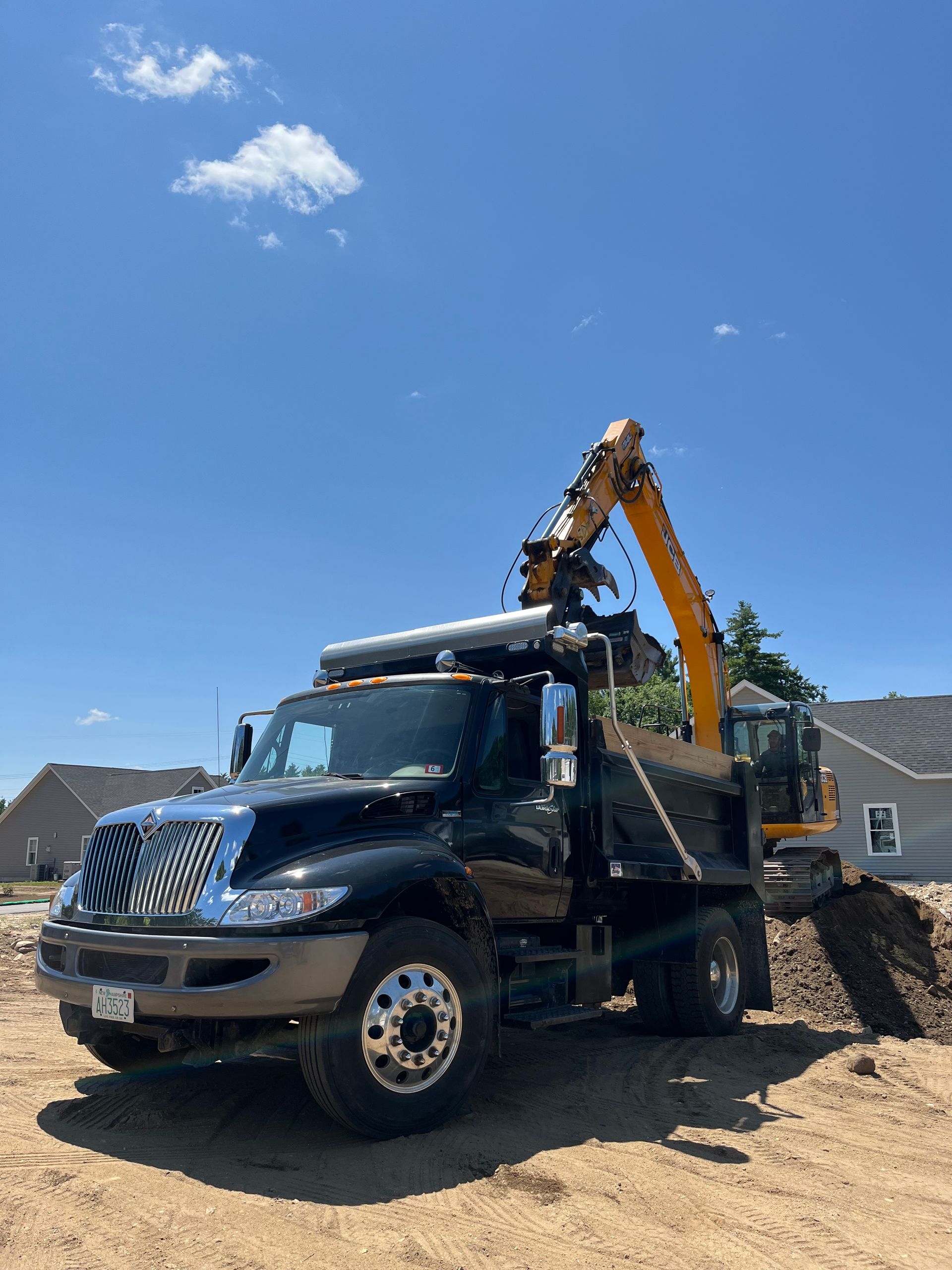 A black dump truck with an attached crane arm loading dirt into its bed at a construction site under a clear blue sky.