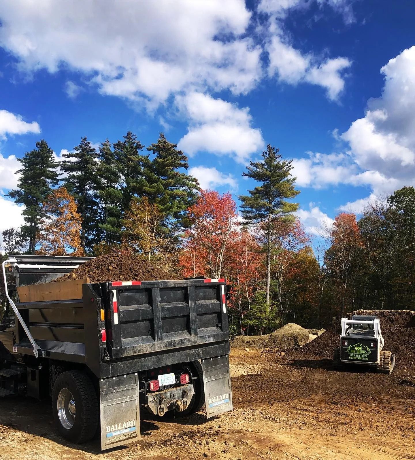 Dump truck and skid steer on a construction site with trees and a blue sky with clouds.
