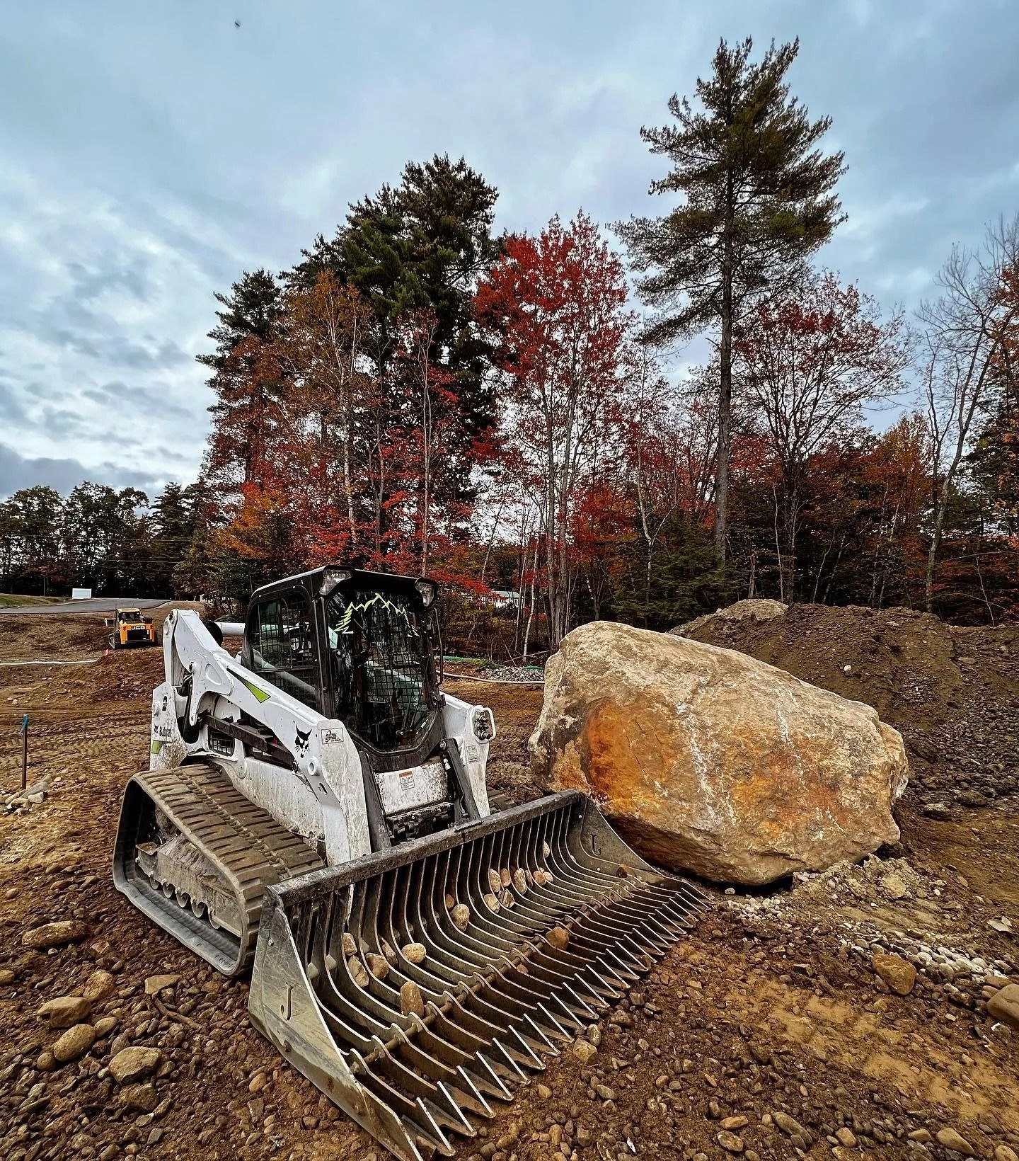 Bobcat skid-steer loader with rock rake next to a large boulder, earth-moving work in progress, fall foliage backdrop.
