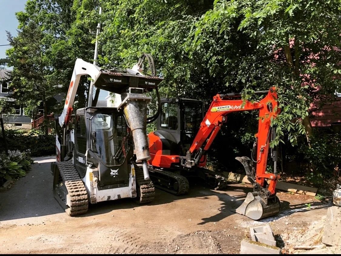 Two construction vehicles: Bobcat with jackhammer and orange excavator on a work site, surrounded by greenery.