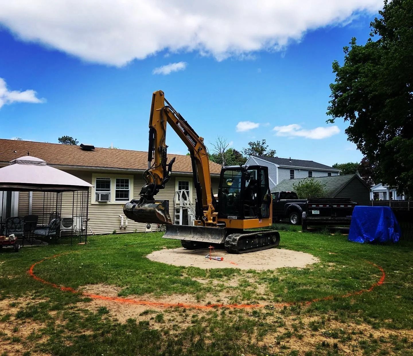 A yellow excavator sits in a backyard on a circular patch of dirt, surrounded by a red spray-painted outline on the grass.