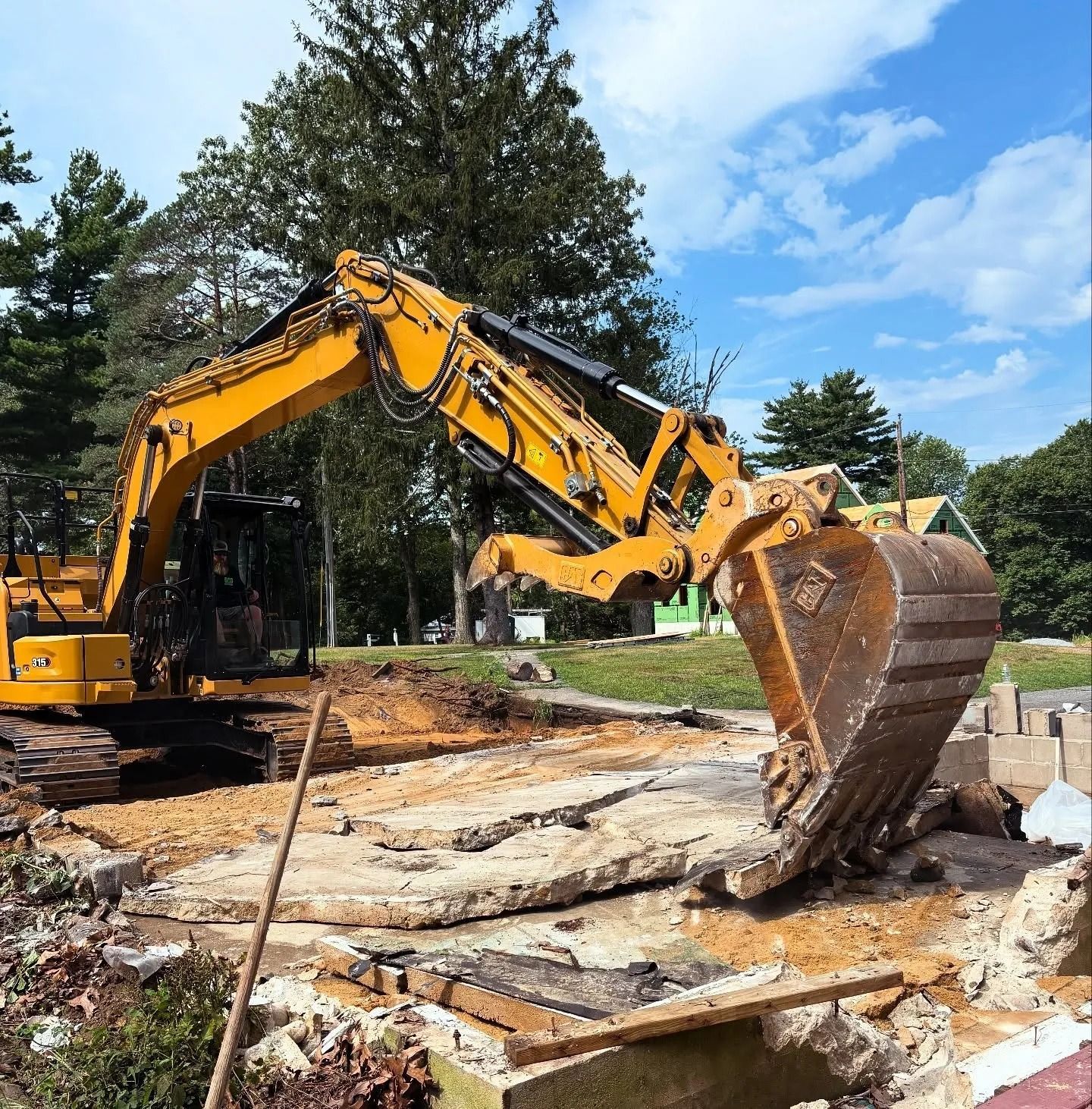 A yellow excavator digging through broken concrete on a construction site with trees and a blue sky in the background.