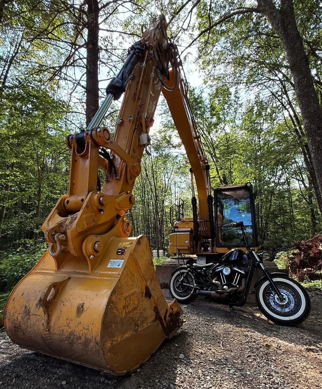 Yellow excavator with black motorcycle parked next to it in a wooded area.