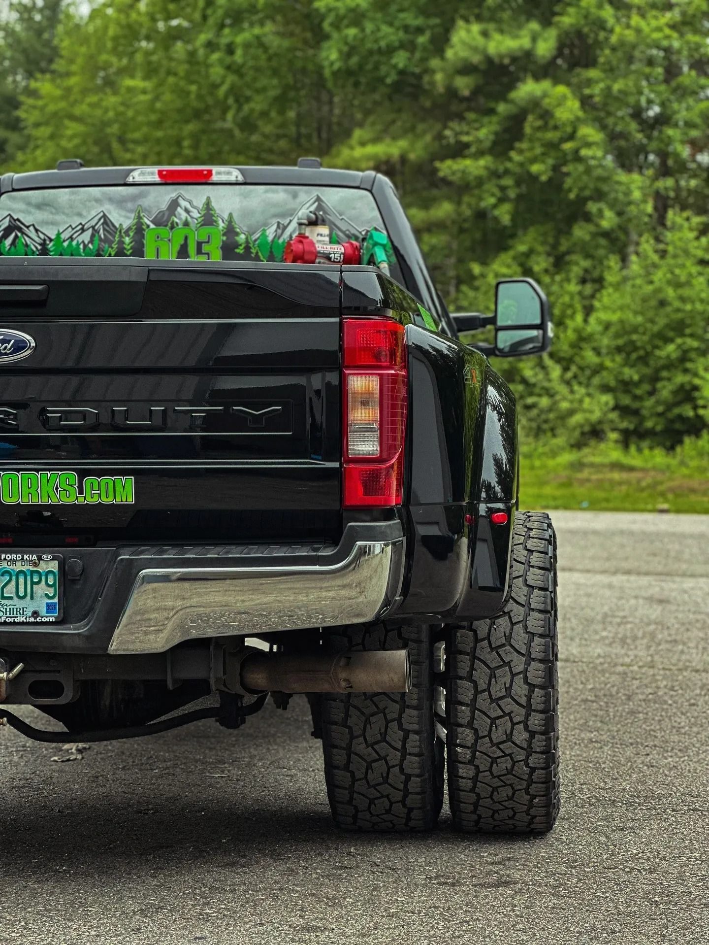 Black Ford dually pickup truck parked on pavement. Rear view, forest in background.