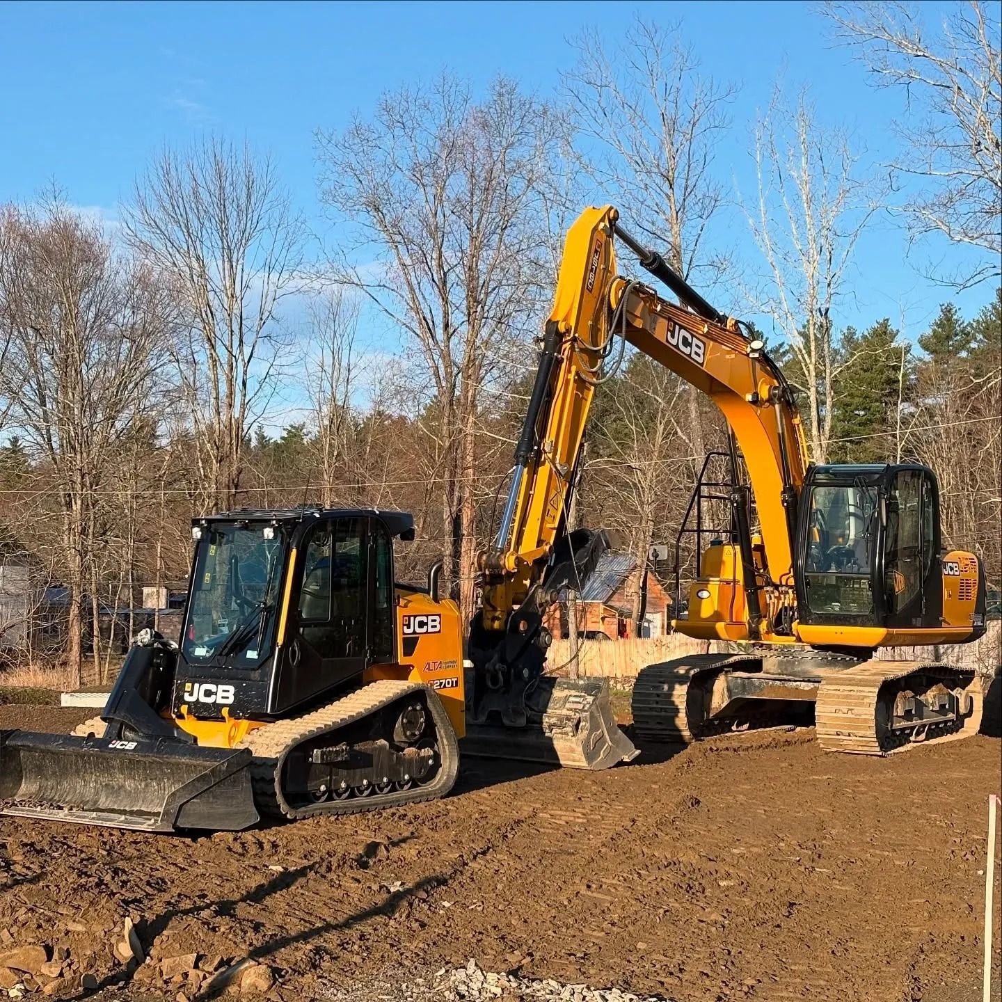 Two JCB construction vehicles on a dirt construction site, one with a front blade, the other a long excavator arm.