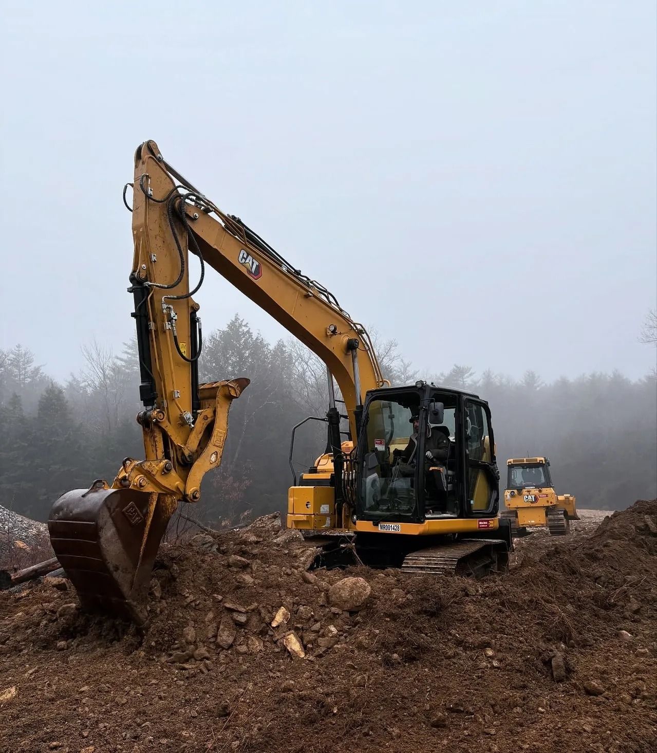 Yellow excavator digging dirt on a cloudy day, with a bulldozer in the background.