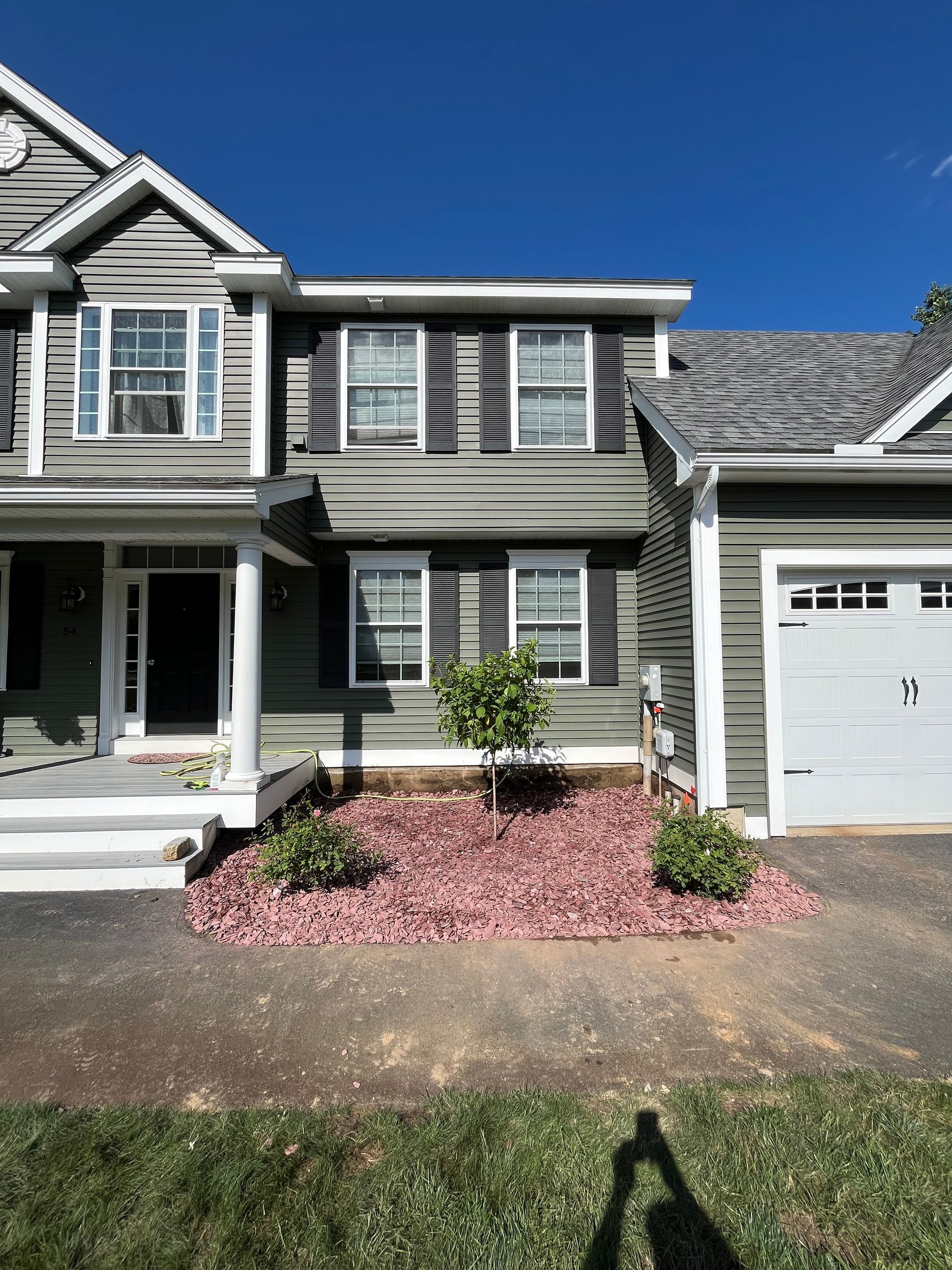A two-story green-sided house with white trim, a front porch, and a white one-car garage beside a red-mulched garden.