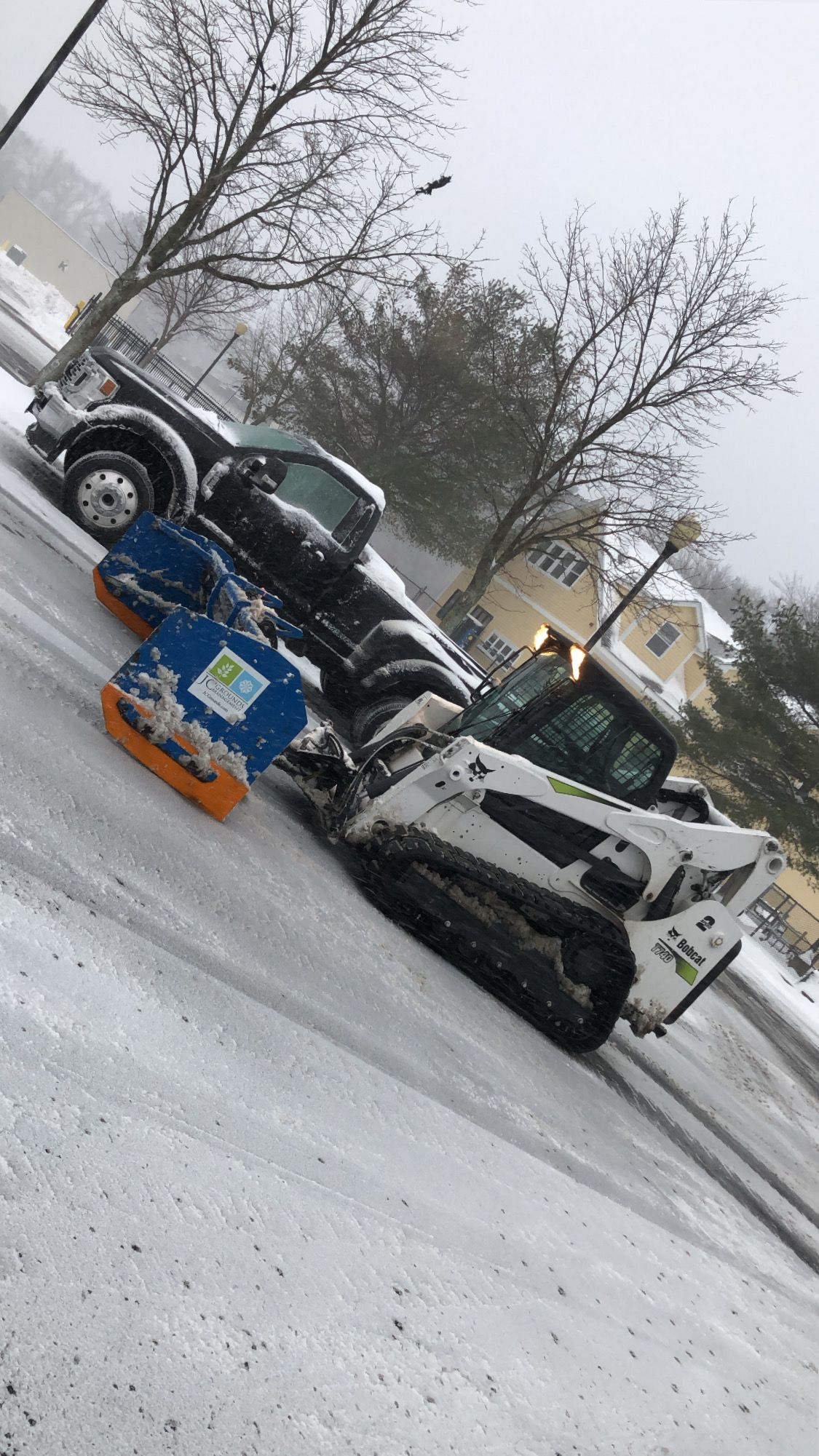 A white Bobcat skid-steer loader with a large snow pusher blade sits on a snowy road near a black pickup truck.
