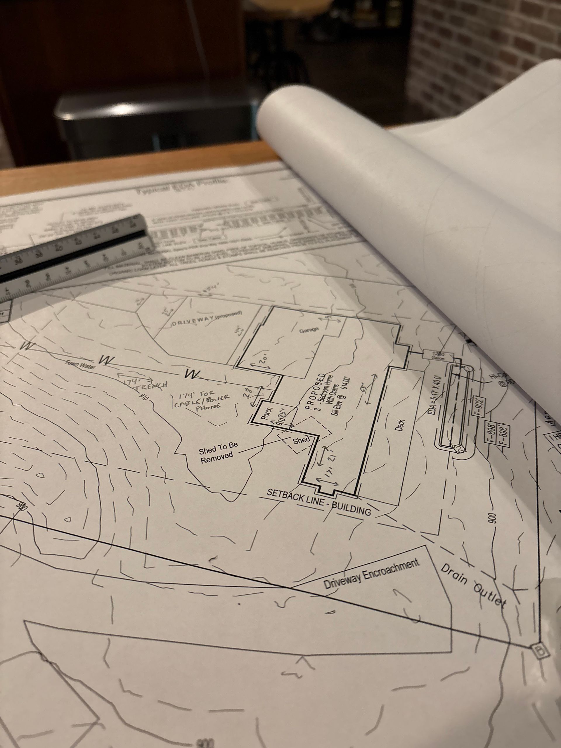 A technical construction or architectural site plan laid out on a table with a metal drafting ruler and rolled paper.
