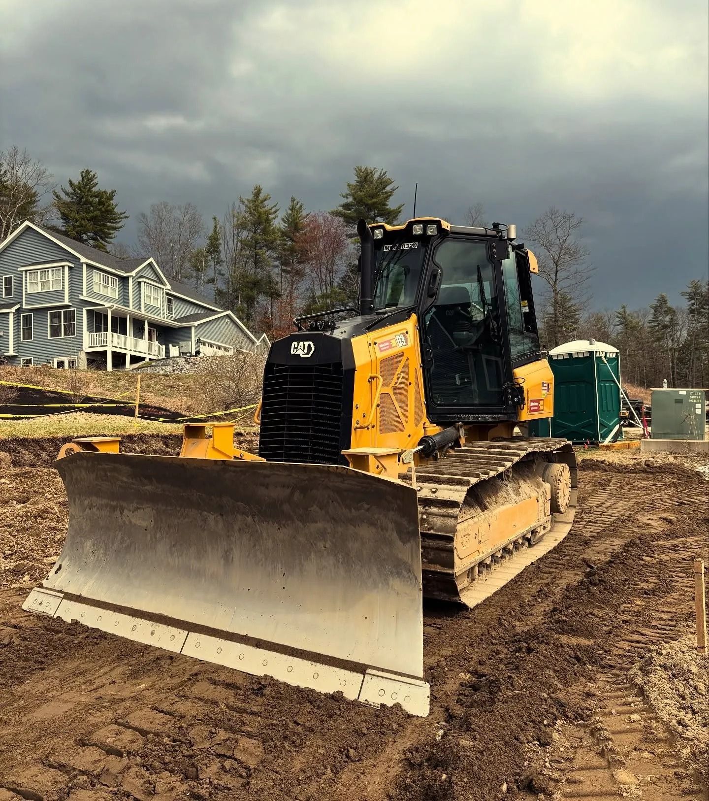 Yellow bulldozer on a muddy construction site, with a house and portable toilet in the background.