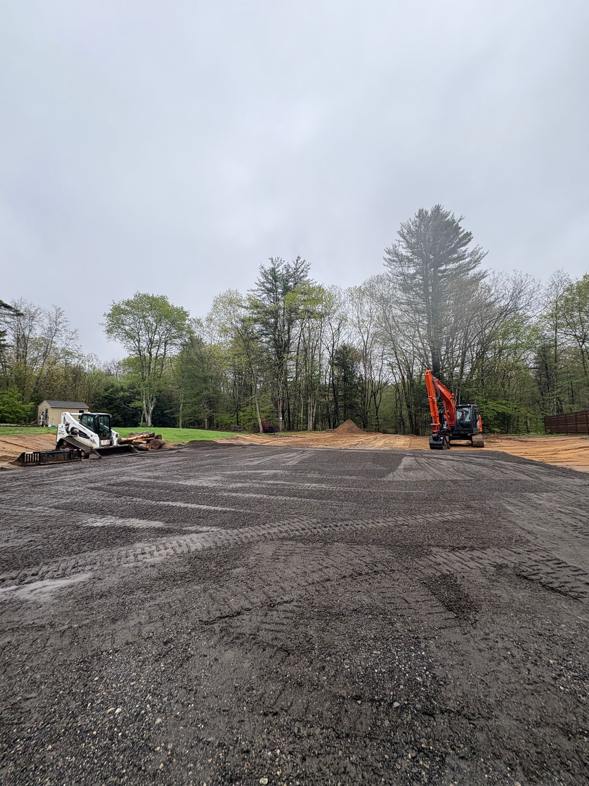 A construction site with a gravel surface, a white skid steer on the left, and an orange excavator against a tree line.