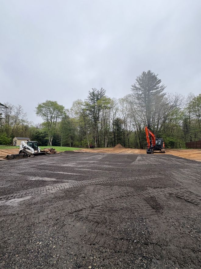 A construction site with a gravel surface, a white skid steer on the left, and an orange excavator against a tree line.