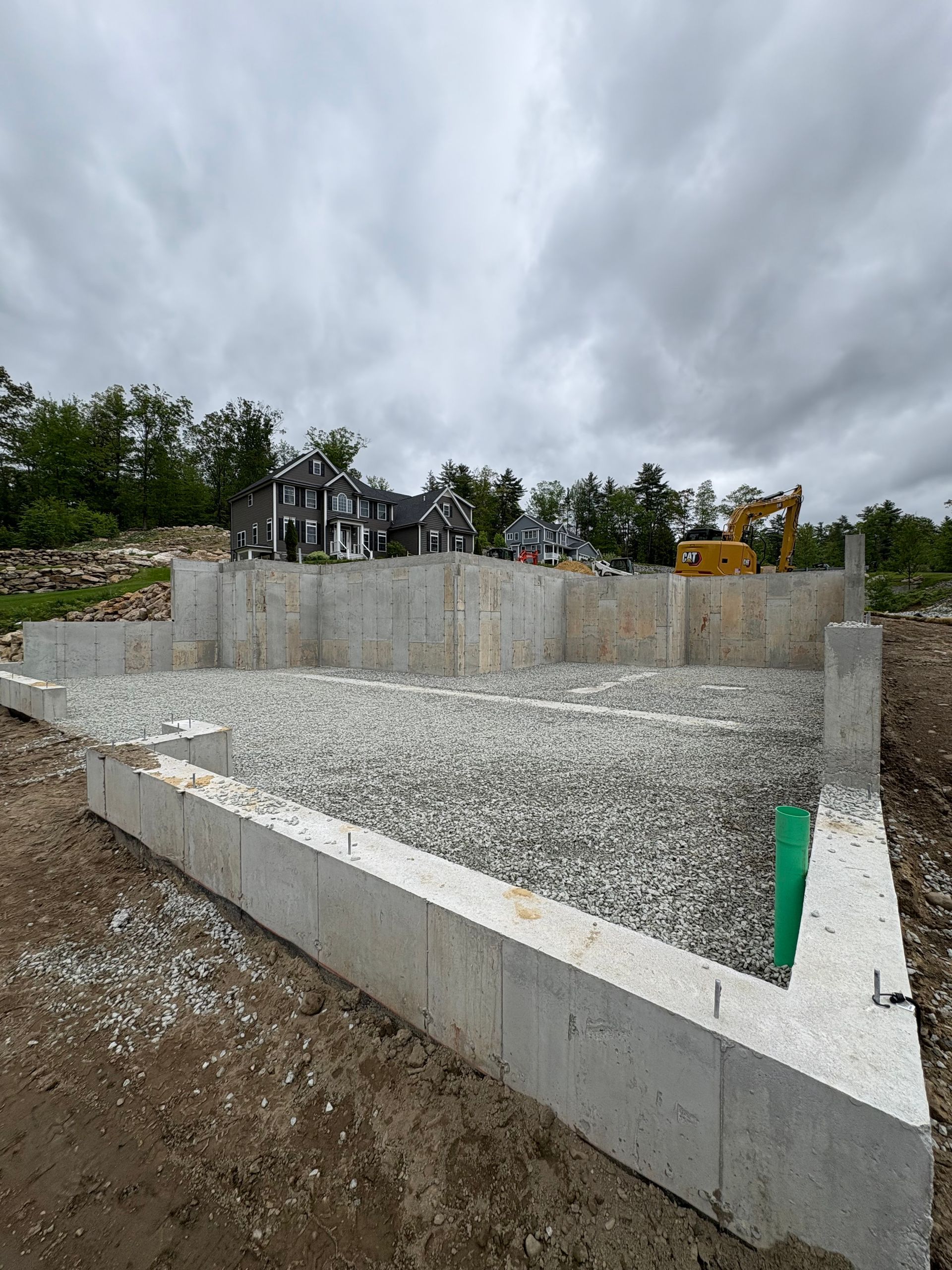 A concrete foundation for a new house under construction, filled with gravel, with a construction vehicle in the distance.