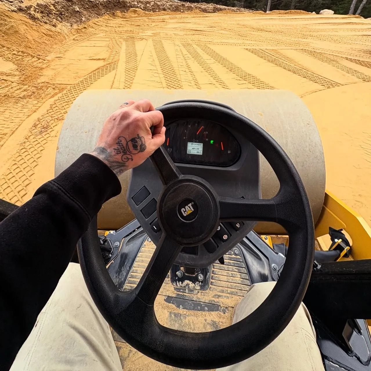 Person driving a Caterpillar compactor on a dirt surface. Steering wheel is in focus.