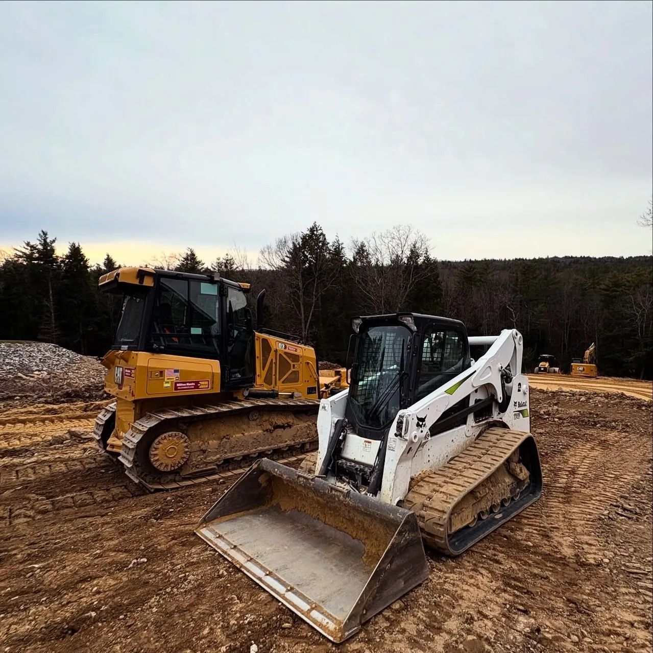 Yellow bulldozer and white Bobcat track loader parked on a construction site. Cloudy sky in background.